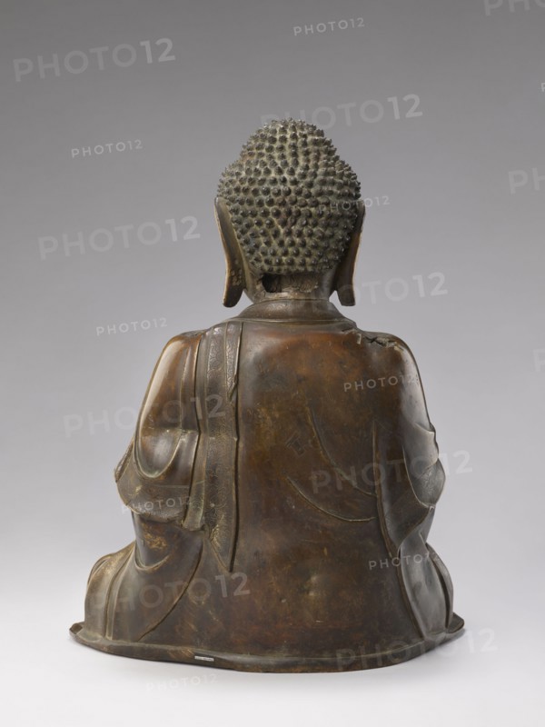 Shakyamuni Buddha, 17th/18th century China, Qing dynasty 1644–1911, Chinese Buddhism; sculpture height 47.0 cm, width 26.0 cm; tutelary deities and Buddha iconography, ushnisha; Chinese art, Qing style., A bronze statue shown from the back depicting a seated figure with a textured hair surface composed of small rounded bumps, elongated ears, and draped robes. The surface colors include dark brown, greenish patina, and hints of lighter brown highlights. The robe displays carved folds and a sash draped over one shoulder with subtle decorative patterns. The base is round and slightly flared, matching the bronze tones of the figure. The background is a smooth gradient of gray.