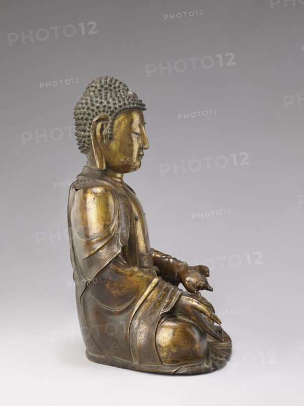 Shakyamuni Buddha, Chinese, Qing dynasty 1644–1911, 17th–18th century, Chinese Buddhism; sculpture of Shakyamuni with ushnisha, tutelary deities and Buddha iconography; height 47.0 cm, width 26.0 cm., A seated statue of a figure in profile facing right, rendered in metallic tones of gold and bronze with areas of darker brown patina; the figure has a textured hair surface of small rounded bumps that appears dark gray to black, a smooth rounded head and elongated earlobes. The figure wears layered robes with decorative borders, showing gradients of gold, bronze, and brown across the folds and rounded shoulders; the hands rest in the lap with one hand slightly cupped above the other, both showing the same metallic gold and brown coloration. The base and lower garments show deeper bronze and brown tones, while highlights on raised areas are brighter gold; the background is a smooth, neutral gray that transitions from lighter at the bottom to darker toward the top.