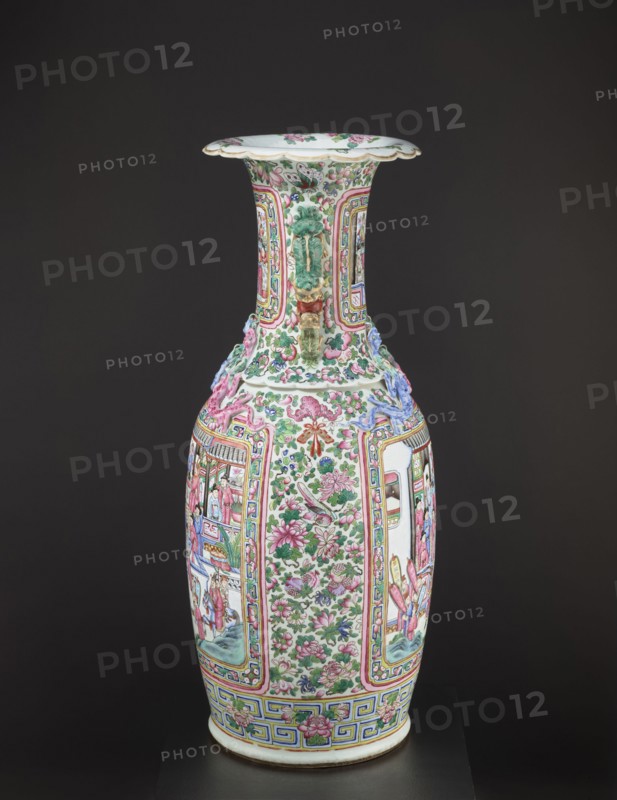 Vase, Chinese, Qing dynasty 1644–1911, colored porcelain, ceramics; subject: lizards, lion iconography, figurative and genre scenes, plant motifs with fruit in the background; decorative vessel/vase; 2nd half of the 19th century; China; height 90 cm, diameter 37.5 cm., A tall porcelain vase with a flared rim and a long neck sits against a dark background; the vase is predominantly white and decorated with a dense pattern of pink and green floral motifs, green leaves, and small birds, with additional colors including blue, yellow, and touches of red and purple; the neck features raised green and gold ornamental handles and panels outlined in pink and yellow, while the body displays alternating vertical panels with intricate scenes and floral fields framed by multicolored borders; a band of geometric patterns in blue and yellow encircles the base, and the rim interior shows scattered floral decoration in pink and green.