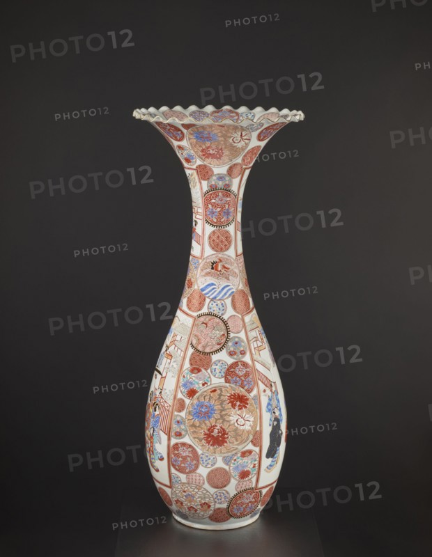 Vase, Japanese; colored porcelain with dragons, figurative scenes, geometric decorations, peonies, chrysanthemums, and other flowers; Meiji period ca. 1870–1880; Arita, Japan; vessel; height 94.0 cm; weight 14.5 kg., A tall, slender porcelain vase with a flared, scalloped rim sits against a dark background; the vase is predominantly white with decorative areas in red, blue, gold, and black forming circular medallions and panels that cover its surface, featuring floral motifs, wave-like patterns, and geometric designs; a few stylized human figures wearing patterned garments appear within some panels; the overall color palette includes white, red, blue, gold, black, and touches of brown, with intricate detailing wrapping the neck and body of the vessel.