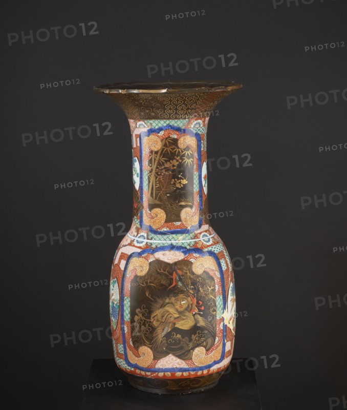 Vase, Japanese, Meiji 1868–1912, c. 1880–1900. High, wide neck with a large flaring cup-like rim; rim with a variegated cut. Decorated with multicolored enamels in the background and in the white reserves; fields covered with black lacquer decorated with gold, red, and gray-blue lacquer; part in flat relief. Motifs: birds, pines, geometric decorations. Colored porcelain vessel, height 79.0 cm, diameter 38.5 cm. Maker and provenance unknown., A tall decorative vase photographed against a dark gray background, predominantly showing colors of black, brown, gold, red, blue, white, green, and orange. The vase has a wide flared rim with a scalloped edge and a dark brown top band decorated with gold swirls. The neck of the vase is narrow and features ornate framed panels with gold-toned scenes on a dark brown background, bordered by patterned bands in blue, red, green, and white with orange scroll motifs. The bulbous lower body of the vase displays a large central panel with a painted figure ...
