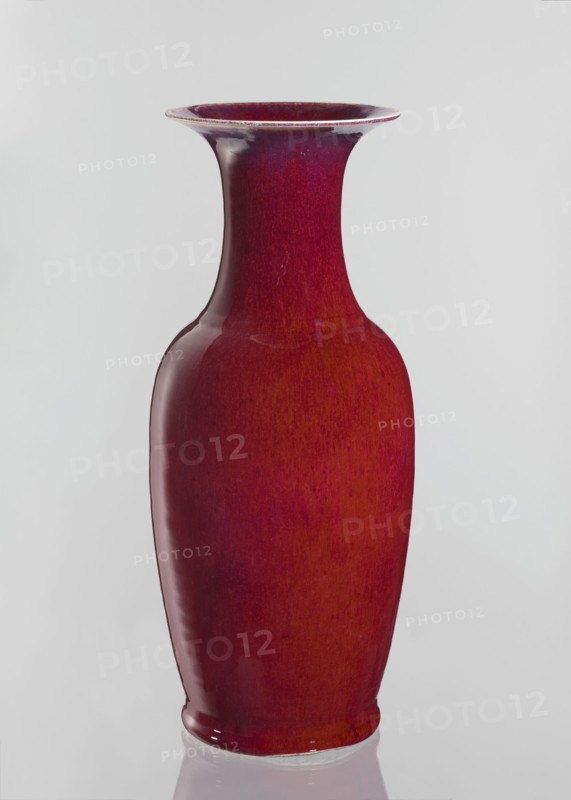 Vase, Chinese, Qing dynasty 1644–1911, 19th century. Large oval ceramic vase with a long wide neck opening, covered with sang de boeuf bull’s blood glaze. Height 58 cm; diameter 24.5 cm., A tall ceramic vase with a flared rim and a narrow neck widening into a rounded body, finished in glossy shades of red and deep crimson with subtle variations and darker red undertones, sitting against a pale gray background and reflected faintly on a glossy surface below.
