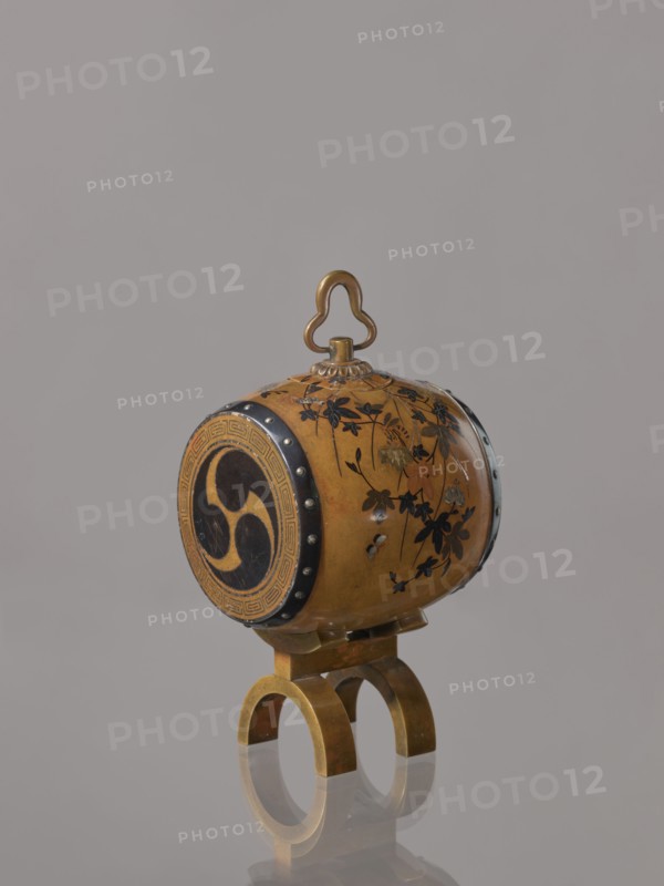 Okimono in the shape of a taiko drum;, possibly Nogawa workshop; ; Japanese, Meiji period ca. 1890–1900; subject: taiko drum with butterflies, insects, plant motifs including paulownia; function: drum-shaped incense burner, vessel; material/technique unspecified; place of origin: Kyoto, Japan; height 15.0 cm, diameter 7.8 cm., An ornate cylindrical object with a rounded top handle sits on a small double-arched stand; the cylinder is primarily golden brown with black floral and leaf decoration painted on its surface, dark circular end panels with a curved triple motif, and darker metal rim accents with small round studs; the stand and handle appear metallic with a brass-like golden color, and the background is a uniform light gray.