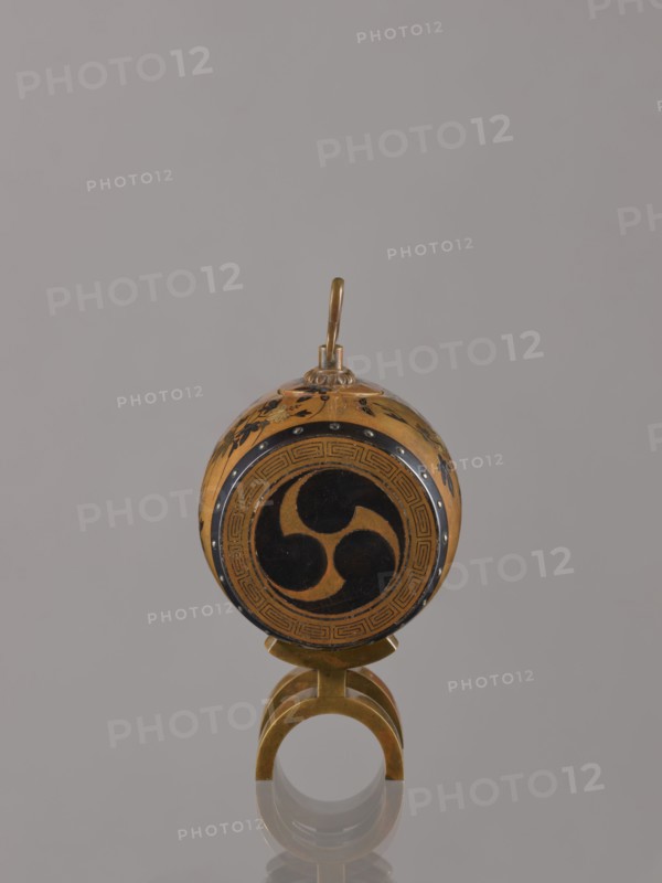 Okimono in the shape of a taiko drum, Nogawa workshop, Japanese, Meiji period ca. 1890–1900. Drum-shaped incense burner, carved with butterflies, insects, and paulownia plant motifs. Kyoto, Japan. Vessel; height 15.0 cm, diameter 7.8 cm., An object with a round, decorated body in shades of gold and black sits upright on a small curved gold stand, featuring a central circular black motif on the front and floral-like black decorations around the sides, with a short gold loop extending from the top; the background is a uniform light gray.