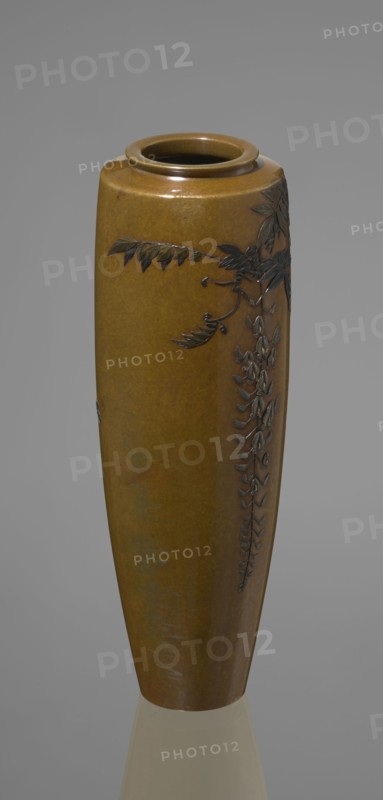 A slender miniature vase by Nogawa, ca. 1890–1900 Meiji period. Silver with gilding and black shakudo patina; round, slightly recessed base with an engraved brand. Belly widens slightly toward the top with narrow, straight arms and a flat-ring spout; no neck. Sparse naturalistic decoration: a wisteria vine placed on the upper part of the belly on one side, with a sumptuous flower cluster hanging down almost to the vase’s end. The masterly combination of various metals and alloys produces a darker-toned decorative motif against the background, giving colorfulness and enhancing the naturalistic yet decorative depiction. Kyoto, Japan. Vessel; height 15.7 cm, width 5.3 cm., A tall vase with a smooth glossy surface in shades of brown and olive green, decorated with a raised dark brown botanical motif of leaves and hanging clusters, set against a neutral gray background and resting on a reflective light surface.