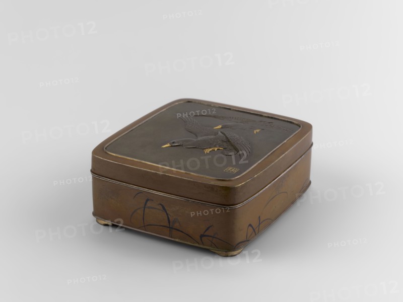 Box with a lid, Jômi, Eisuke I 1839-1899 author. Rectangular box with rounded corners, decorated on the flat lid with two flying wild geese made in high relief with inlay of silver and gold iro-e zogan technique. The side walls are decorated with a motif of inclined reeds, made of silver and shakudo using the hira-zogan technique, so the decorative motif is level with the background surface, giving a painted effect. On the lid there is a signature made in gold: in a square field, two stylized kanji characters resembling archaic ones: Shobi. ca. 1880–1899 Meiji Period, 1868–1912, Kyoto, Japan. Container. Height 4.8 cm; width 10.2 cm; depth 11.4 cm., A rectangular metal box with rounded corners sits on a neutral light gray surface; the box is mainly brown with darker brown and black areas, and small accents of gold on a relief of birds on the lid. The lid features a raised design of a group of birds in flight with textured wings and gold-tipped beaks and feet, and the sides of the box sh...