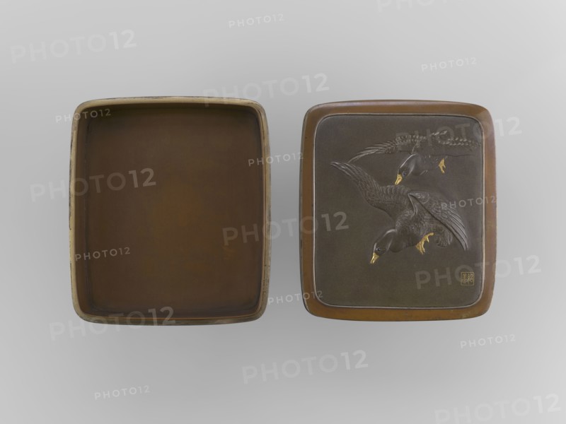 Box with a lid, Jômi, Eisuke I 1839–1899 artist; . Rectangular box with rounded corners, decorated on the flat lid with two flying wild geese made in high relief with inlay of silver and gold iro-e zogan technique. The side walls are decorated with a motif of inclined reeds, made of silver and shakudo using the hira-zogan technique, where the decorative motif is level with the background surface producing a painted effect. On the lid there is a signature in gold in a square field of two stylized kanji characters resembling archaic forms: Shobi. Ca. 1880–1899 Meiji Period, 1868–1912, Kyoto, Japan. Container. Height 4.8 cm; width 10.2 cm; depth 11.4 cm., A pair of rectangular trays displayed side by side on a light gray background, one tray shown open with a smooth brown interior and slightly worn outer rim with hints of beige, the other tray closed with a darker brown surface featuring a raised design of a group of birds in flight with detailed feathers and golden beaks and feet, the cl...