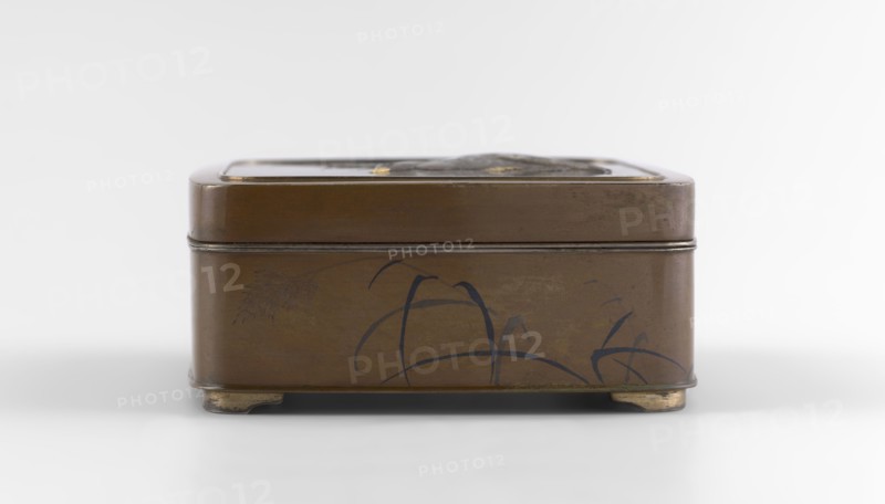 Box with a lid, Jômi, Eisuke I 1839–1899 artist; rectangular box with rounded corners, decorated on the flat lid with two flying wild geese made in high relief with inlay of silver and gold iro-e zogan technique. The side walls are decorated with a motif of inclined reeds, made of silver and shakudo using the hira-zogan technique, producing a painted effect. The lid bears a gold signature in a square field with two stylized kanji characters resembling archaic forms: Shobi. Ca. 1880–1899 Meiji period, 1868–1912. Japan, Kyoto. Container. Height 4.8 cm; width 10.2 cm; depth 11.4 cm., A small rectangular metal box with rounded corners and a lid sits on a plain light surface against a pale background; the box is predominantly brown with darker brown edges and faint darker markings on its sides, featuring a few curved dark blue brushlike strokes and a subtle darker brown leaflike motif, and it rests on small worn metallic feet that show a golden-beige tone.