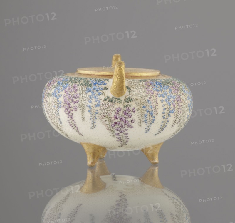 Incense burner koro with cover by Kinkozan Sobei VII 1867–1927, Kinkozan workshop; ceramics Kyoto satsuma, Meiji period 19th/20th century. Earthenware censers/koro; three-legged vessel with two handles, decorated with wisteria, chrysanthemums and flowers; signed. Height 7.8 cm; diameter 9.5 cm. Japan: Kyoto, Awata., A rounded ceramic vessel with a pale cream background sits on three short gold-colored legs and has a gold-colored rim and handle; the body is decorated all around with hanging clusters of small flowers in shades of purple, blue, and pink with green leaves, and the surface shows a fine dotted texture; the gold-colored legs and handle have subtle floral embossing, and the vessel and its legs are reflected on a smooth gray surface beneath; the background is a uniform muted gray.
