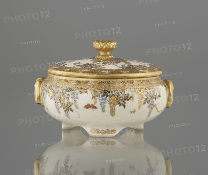 Small box with a lid, Kinkôzan, Sôbei VII 1867–1927 author, Kinkôzan workshop / co-creator; Japanese, Meiji late 19th century; Kyoto, Awata. Satsuma ceramics vessel decorated with wisteria and butterflies. The lid has a gold knob in the shape of a flower. The vessel has two slightly convex handles on three wide feet. Signed. Height 6.8 cm; diameter 10.2 cm., A small lidded porcelain bowl with a rounded body and short feet sits on a reflective surface against a gray background; the bowl is primarily white with ornate gold trim and gilded handles on each side, and the lid has a gold knop; delicate painted floral garlands in colors including blue, pink, green, purple, and brown hang around the body and lid, with small painted insects in red and yellow visible among the flowers; the reflection of the bowl and its decoration is visible on the surface below.