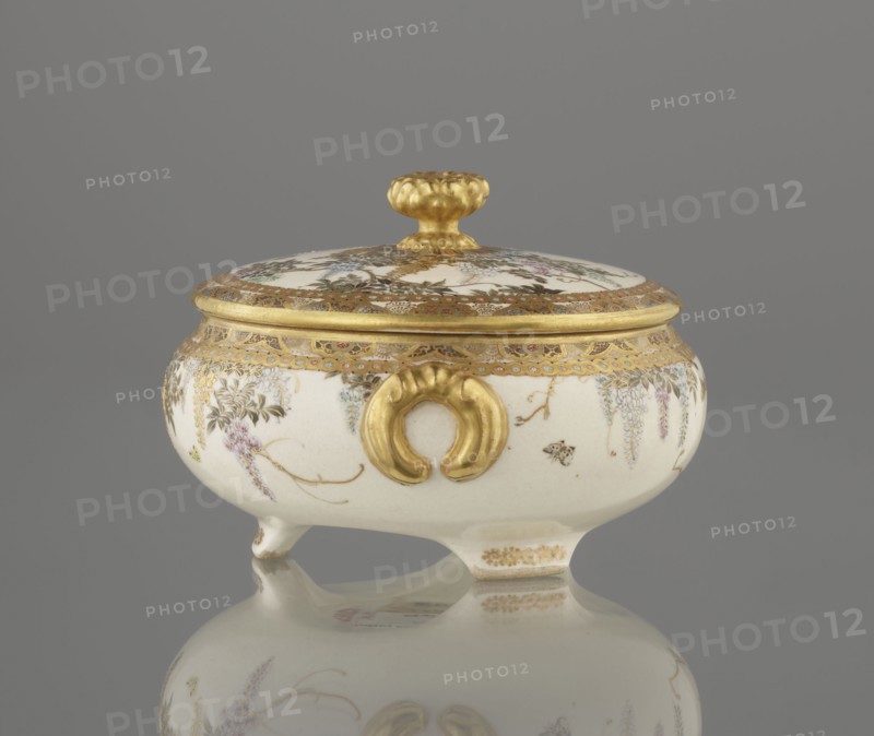 Small box with a lid, Kinkôzan, Sôbei VII 1867–1927 author, Kinkôzan workshop/co-creator; ; Japanese art; Japanese; Meiji 1868–1912; wisteria; ceramics; butterflies; boxes; satsuma. The lid with a gold knob in the shape of a flower. The vessel with two slightly convex handles, on three wide feet. Signed, late 19th century Meiji period. Japan; Kyoto, Awata. Vessel, container; height 6.8 cm; diameter 10.2 cm., A small lidded ceramic vessel with a rounded white body and a matching domed lid, both decorated with delicate painted floral motifs in muted purples, greens, and pale blues, and detailed gold-painted borders and accents; the lid has a textured gold knob, and the vessel's body features a prominent gold ornamental handle on the front and several small gold decorative trims, all resting on short feet and reflected on a smooth gray surface, with the background a uniform gray.