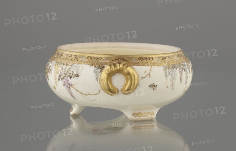 Small box with a lid, Kinkôzan, Sôbei VII 1867–1927 maker; Kinkôzan workshop. . Japanese, Meiji period late 19th century. Ceramics satsuma. Kyoto, Awata. The lid has a gold knob in the shape of a flower. The vessel has two slightly convex handles and rests on three wide feet. Signed. Subject motifs: wisteria and butterflies. Vessel/container. Height 6.8 cm; diameter 10.2 cm., A low, rounded ceramic bowl with a glossy white surface sits on small feet and casts a reflection on a smooth gray surface, decorated with gold ornamental trim around the rim and a prominent raised gold handle-like motif on the side, with delicate painted floral garlands and hanging blossoms in soft purple and pale blue tones accented by fine brown stems and green leaves, and small touches of brown in tiny insect-like details against a neutral beige interior and a muted gray background.