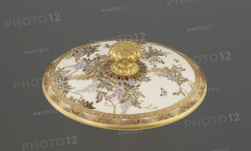 Small box with a lid, Kinkôzan, Sôbei VII 1867-1927 author; Kinkôzan workshop/co-creator; wisteria, ceramics, butterflies, boxes, satsuma. The lid with a gold knob in the shape of a flower. The vessel with two slightly convex handles, on three wide feet. Signed, late 19th century Meiji period, Japan, Kyoto, Awata. Vessel/container; height 6.8 cm, diameter 10.2 cm., A round decorative lid with a central raised knob sits against a gray background; the knob and outer rim are gold, and the flat surface is primarily white with ornate floral and foliate patterns in gold, green, blue, pink, and brown, plus a few small butterfly-like motifs; a colorful radial motif in red, green, and blue surrounds the base of the knob, and a gold patterned border runs around the edge.