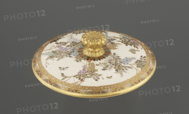 Small box with a lid, Kinkôzan Sôbei VII 1867–1927 maker, Kinkôzan workshop, Japanese, Meiji period late 19th century, Kyoto, Awata. Satsuma ceramics vessel/container with wisteria and butterflies decoration. The lid has a gold knob shaped like a flower. The vessel has two slightly convex handles and stands on three wide feet. Signed. Height 6.8 cm; diameter 10.2 cm., An oval lid with a domed golden knob sits against a gray background, the lid surface predominantly creamy white with ornate botanical and insect illustrations in shades of gold, brown, green, blue, pink, and purple, surrounded by a wide decorative gold border featuring patterned accents, and the central knob rests on a multicolored sunburst-like base in red, blue, green, and gold.