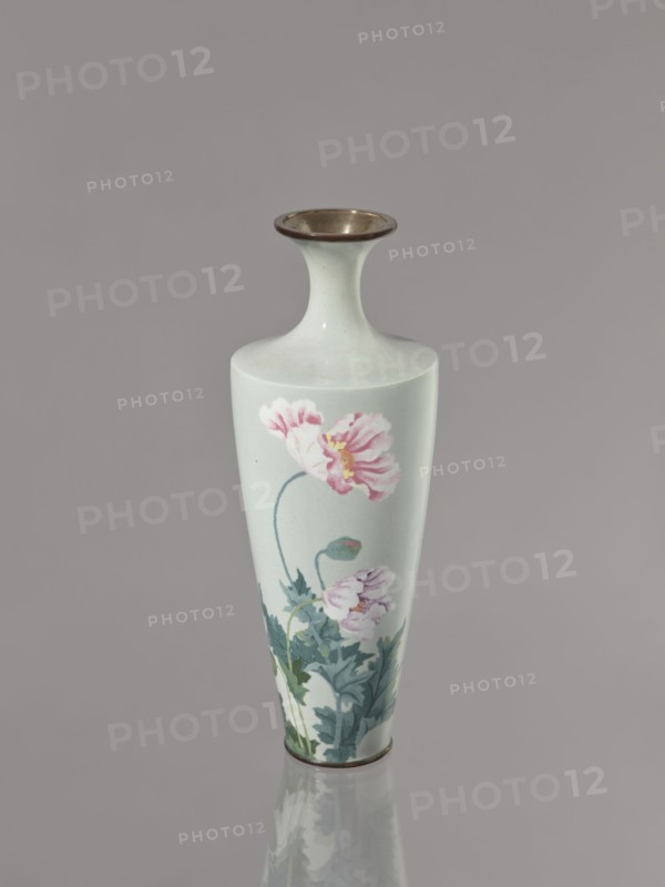 Vase, gift, cloisonné enamel, Symmetrical decoration: blooming poppies on a grey-blue background; flowers poppies. Meiji–Taisho period, ca. 1910–1920. Nagoya, Japan. Vessel; height 24.5 cm, diameter 9.0 cm., A tall, slender vase with a flared neck and rounded lip sits against a plain background. The vase is a pale, light greenish-white color with a glossy surface and a metallic rim at the top and base. Painted on its body are delicate floral motifs: a few large pink flowers with layered petals and pale yellow centers, accompanied by stems and a cluster of bluish-green leaves at the lower portion. The vase casts a soft reflection on the surface beneath it. The visible colors include pale greenish-white, pink, yellow, bluish-green, metallic bronze, and muted gray from the background and reflection.