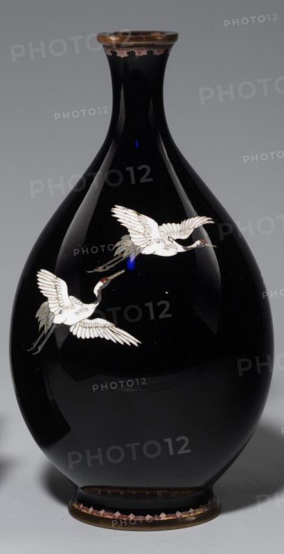 Vase, gift; cloisonné enamel; Japanese; Meiji period 1868–1912, 19th/20th century; Nagoya, Japan; subject: cranes iconography; With a thin, short neck widening towards the spout; vessel; height 18.3 cm, diameter 10 cm., A glossy black vase with a rounded body and a narrow neck, featuring a metallic rim at the top and a metallic base trim at the bottom; the body shows a decorative motif of a few white cranes in flight with pale gray and muted green tail feathers and small red accents on their heads, and there are subtle reflections and highlights on the vase surface showing deep black and dark gray tones against a neutral gray background.