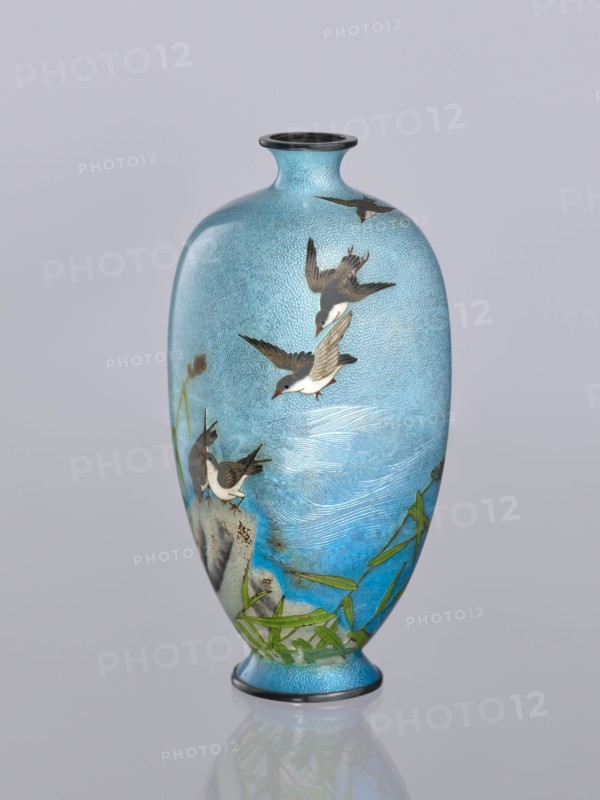 Vase, Tomiki, Shôbei fl. ca. 19th/20th century craftsman, chidori plovers, cloisonné enamel, Japanese art, reeds, waves, decorative vessel, Meiji 1868–1912, Signature: Tomiki engraved on the bottom in a vase-shaped cartouche, 3 katakana characters: Tomiki; artist Tomiki Sobei, 19th/20th century Meiji period, Japan; vessel; height 12.2 cm, width 5.6 cm, depth 5.6 cm, A tall, rounded vase with a narrow neck and flared rim sits against a pale gray background; the vase is predominantly light blue with a textured, speckled surface and areas of deeper blue, green, brown, white, black, and gray. The body of the vase is decorated with a scene of several birds in various flying and perched poses, their feathers rendered in shades of brown, white, black, and gray with small red accents on their beaks or feet. Near the base and lower sides there are stylized green leaves and stems, and a rocky motif in gray and brown. Subtle wave-like markings in lighter blue and white suggest water across the mi...