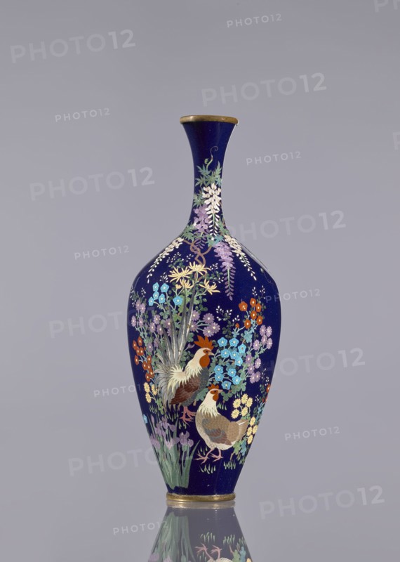 Vase, cloisonné enamel, wisteria, hens, flowers, roosters iconography, Japanese art, Meiji 1868–1912, ovoid vase elongated with a long thin neck slightly flared at the spout, navy blue background of the decoration, c. 1890–1910, Japan, vessel, height 15.6 cm, dia. 5.6 cm, width 6.4 cm, A tall vase with a narrow neck and rounded body sits against a plain light gray background, its surface a deep navy blue adorned with a detailed floral and bird design in colors including purple, lavender, light blue, turquoise, orange, yellow, green, brown, white, and gold; the painted scene shows a pair of birds with brown and white feathers among clusters of various flowers and long green leaves, with cascading floral elements near the neck and a gold rim at the top and base, and a reflection of the vase visible on the glossy surface beneath.
