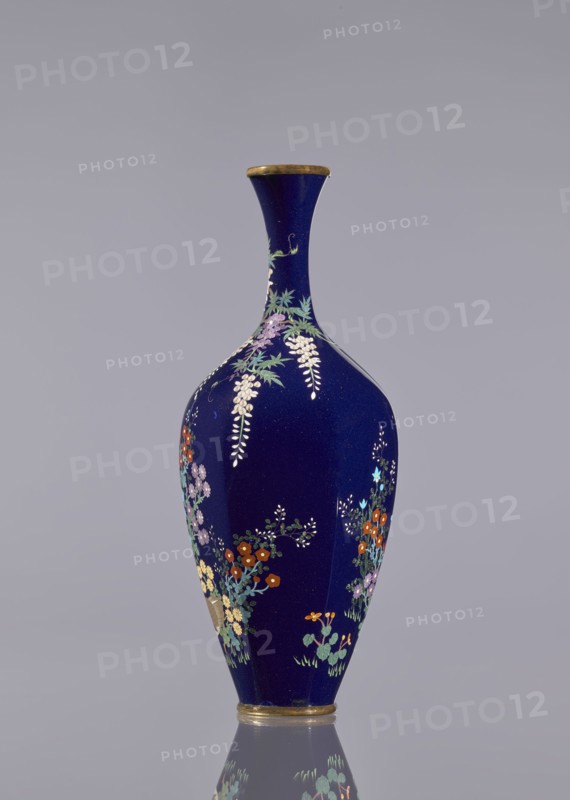 Vase, cloisonné enamel, wisteria, hens, flowers, roosters iconography, Japanese art, Meiji 1868–1912, Ovoid vase, elongated, with a long thin neck slightly flared at the spout, navy blue background of the decoration, c. 1890–1910, Japan, vessel, height 15.6 cm, dia. 5.6 cm, width 6.4 cm., A tall, slender vase with a deep blue glossy surface stands against a plain light gray background, its narrow neck flaring slightly at the top with a thin metallic rim and a matching metallic base. The body of the vase is decorated with a group of delicate floral motifs in colors including white, purple, pale yellow, orange, red, light blue, green, and turquoise, with hanging clusters of pale blossoms and leafy sprigs arranged around the curve. Reflections at the base show a faint mirrored image of the lower floral decorations and the blue color.