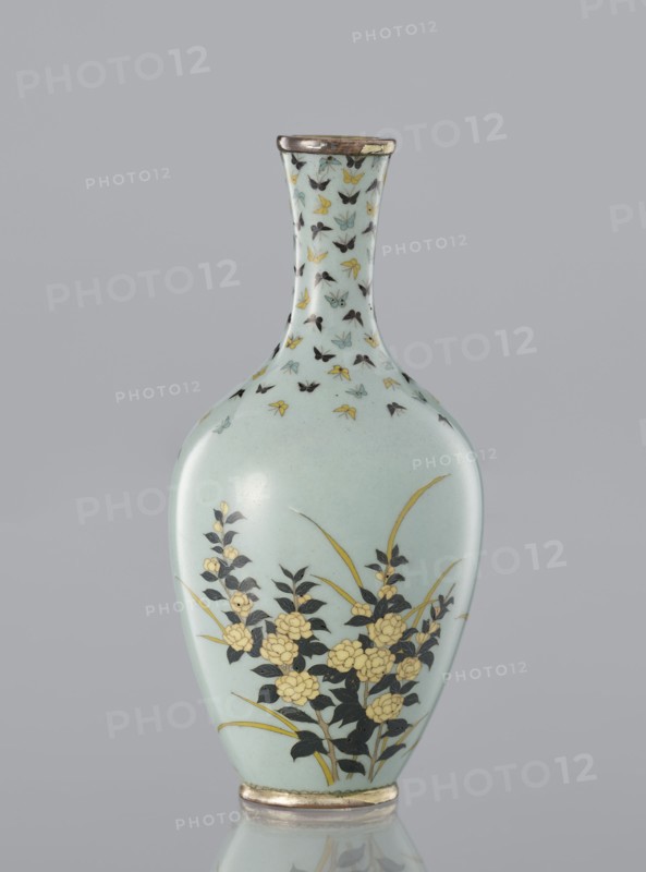 Vase with a thin, very slightly flared neck. Decoration of flowers and butterflies in cloisonné enamel on a pale blue-gray background. Japanese, ca. 1910–1920 Meiji/Taisho periods. Material: cloisonné enamel. Height 11 cm; diameter 5.3 cm., A pale blue ceramic vase with a narrow neck and rounded body decorated with a repeating pattern of small stylized butterflies in black, yellow, and light blue around the neck, and a floral motif on the body showing dark leaves and stems with clusters of yellow blossoms and a few long yellow-green leaves; the rim and base have metallic-looking bands in a worn silver or bronze tone, and the vase sits against a neutral gray background.