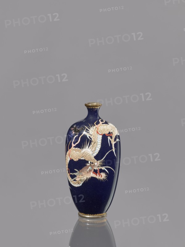 Vase, Japanese, Meiji 1868–1912, cloisonné enamel, 19th/20th century, depicts Japanese dragon motif/iconography, vessel, height 36.7 cm, diameter 4.6 cm., A dark blue ceramic vase with a narrow neck and flared rim sits upright against a gray background, featuring a decorative dragon motif in shades of cream, beige, red, and gold that winds around its body; the rim and base have gold-colored accents, and the vase casts a subtle reflection on the surface below.