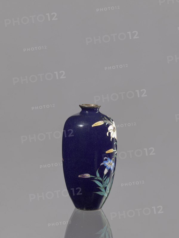 Vase, gift, repossession Krakow, lilies, Japanese art, Japanese, Meiji 1868–1912, cloisonné enamel, 19th/20th century Meiji period, Japan, vessel, height 9.1 cm, diameter 4.8 cm, A tall vase with a deep blue glossy surface stands on a reflective surface, showing a partial reflection below. The vase has a narrow neck with a slightly flared rim in a metallic color and a rounded body that tapers toward the base. On one side of the vase there is a painted floral design featuring a few white flowers, a few blue flowers, several yellow buds, and green leaves with visible stems. The background behind the vase is a uniform light gray.