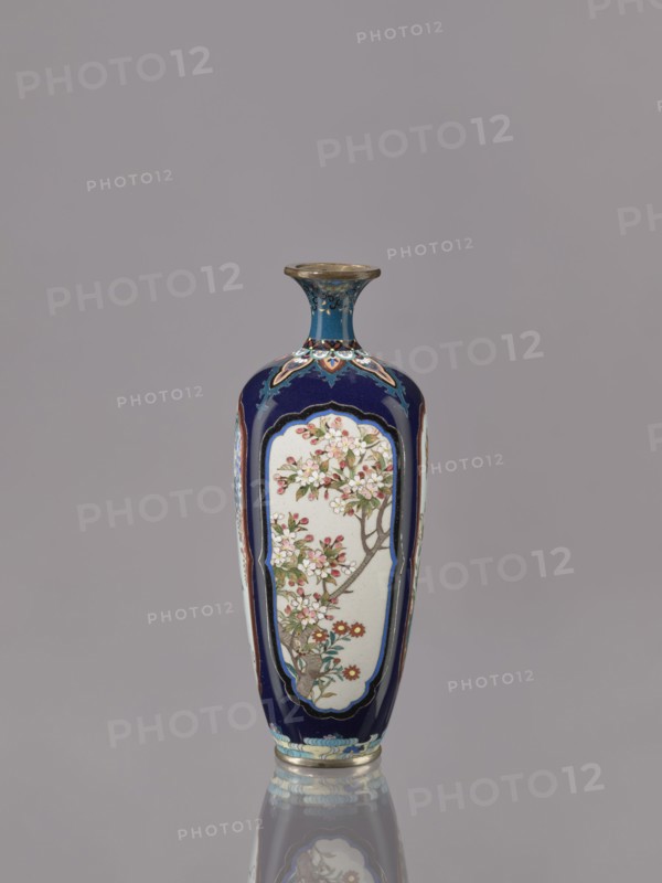 Vase, Ovoid elongated cloisonné enamel vase with a short, thin, strongly flared neck. Decoration: four reserves depicting blossoming cherry, apple tree, wisteria, and lilies on a navy blue background. Japanese; Meiji–Taisho period, ca. 1910–1920. Vessel; height 15.5 cm, diameter 6.0 cm., A tall, narrow vase with a flared neck sits against a plain gray background, predominantly dark blue with panels of off-white featuring floral branch motifs in shades of pink, red, green, brown, and white, with gold and light blue decorative borders and accents, and a metallic rim and base showing reflections on the surface below.