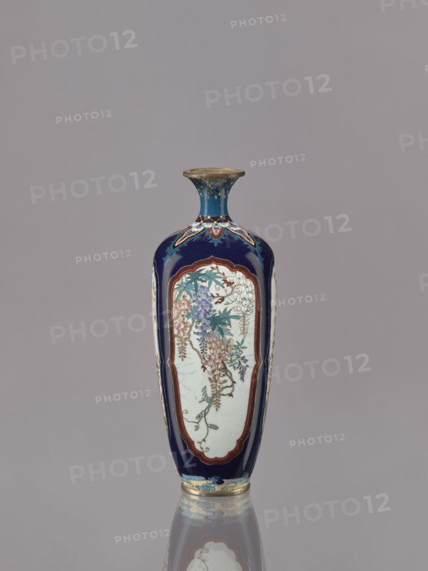 Vase, cloisonné enamel, ovoid elongated form with a short, thin, strongly flared neck. Decoration in four reserves depicting blossoming cherry, apple tree, wisteria, and lilies on a navy blue background. Japanese, ca. 1910–1920 Meiji/Taisho periods. Vessel; height 15.5 cm, diameter 6.0 cm., A tall, narrow vase with a flared rim stands against a neutral gray background, predominantly featuring deep blue and white surfaces with accents of gold, teal, purple, pink, green, and brown; the body displays elongated white panels framed in brown on a dark blue ground, each panel decorated with cascading floral clusters and leafy stems in shades of purple, pink, teal, and green, with delicate outlining and small brown branches; the neck is blue with gold and teal decorative motifs and a gold rim, while the base has decorative bands in blue, teal, and gold; the vase casts a subtle reflection on the surface below.