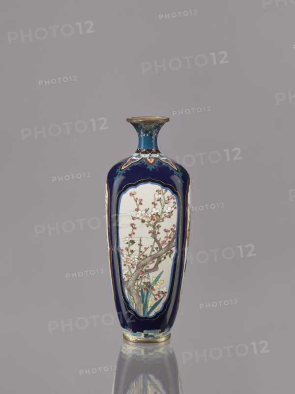 Vase, Ovoid elongated cloisonné enamel vase with a short, thin, strongly flared neck. Decoration: four reserves depicting blossoming cherry, apple tree, wisteria, and lilies on a navy blue background. Japan, ca. 1910–1920 Meiji/Taisho periods. Vessel; height 15.5 cm, diameter 6.0 cm., A tall, narrow vase with a flared rim sits upright against a plain gray background, predominantly colored deep blue with panels of pale off-white featuring a delicate floral design showing branches and blossoms in shades of pink and white with green leaves and brownish-gray stems; the vase also includes accents of gold, turquoise, red, and yellow in decorative borders and small painted details, and the surface reflects faintly below creating a subtle mirrored effect.