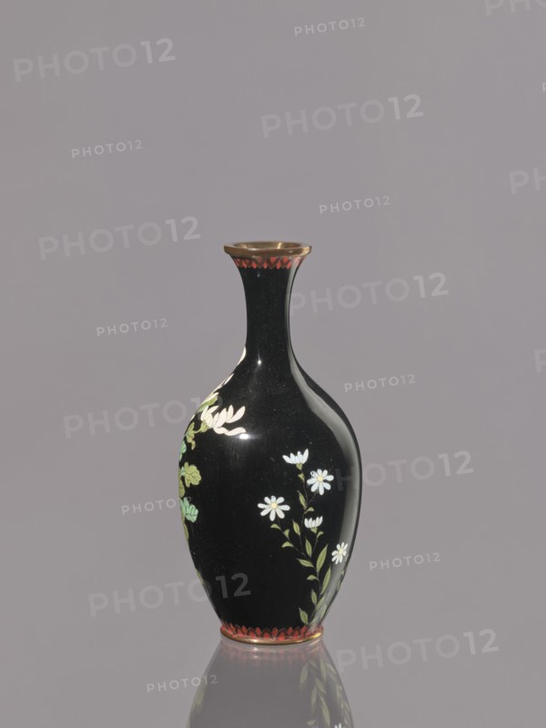 Vase, chrysanthemums, Japanese art, Japanese culture, flowers iconography, Meiji 1868–1912, cloisonné enamel, symmetrical decoration, ca. 1900, Nagoya, Japan, vessel; height 19.3 cm, diameter 9.1 cm., A glossy black vase with a narrow neck and flared rim stands against a muted gray background, decorated with painted white flowers and green leaves along its rounded body, a band of red decorative scallops near the rim and base, and a reflective surface that creates a subtle mirrored reflection below.