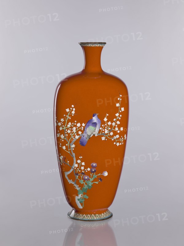 Vase, cloisonné enamel; decorated with chrysanthemums, cherries, and birds; Meiji–Taisho period, ca. 1910–1920; Nagoya, Japan; height 24.0 cm, width 10.4 cm, depth 8.5 cm., An orange vase with a narrow neck and flared rim stands against a pale gray background, decorated with a painted branch bearing small white blossoms and green leaves; a purple and blue bird perches on the branch, and a few purple flowers appear near the base, while the rim and foot have subtle patterned bands in muted green and cream.