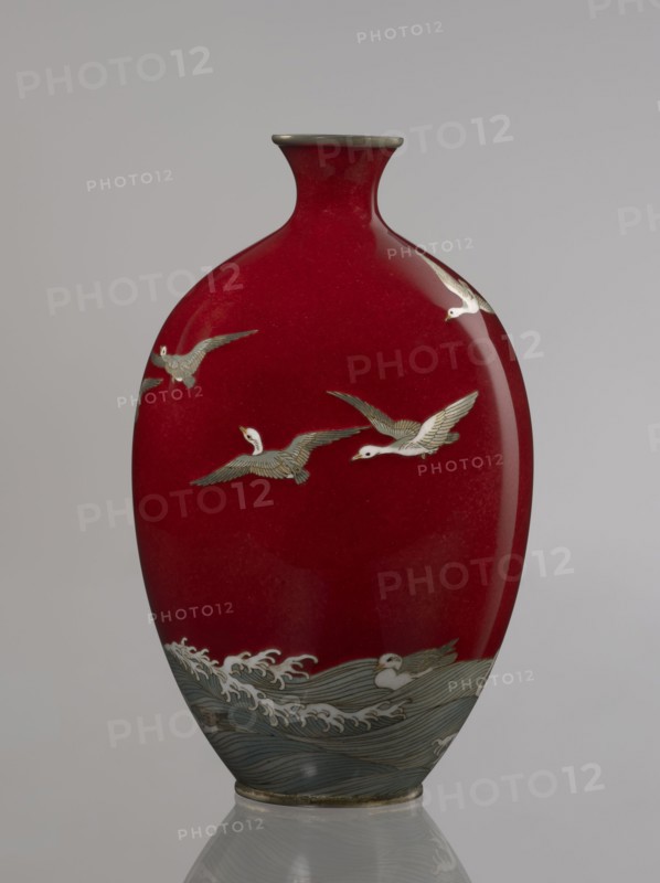 Vase by Hayakawa Komejirô fl. ca. 1880–1912?, late 19th century Meiji period, 1868–1912, Nagoya, Japan. Small ovoid, bulging bottle on a very low round foot framed in silver-plated metal, with a short narrow neck and flared spout also framed in silver-plated metal. Body covered with transparent red enamel akasuke technique, developed around 1880 in Nagoya. Motif of water birds—plovers chidori—executed with the yusen technique using thin silver wires, shown flying over foaming waves or perching on the water; design combines naturalism and decorative stylization typical of Japanese design. Signature engraved in kanji on the bottom: Hayakawa zo tsukuru. Vessel; height 16.7 cm, diameter 9.9 cm., A glossy vase with a deep red body and a narrow metallic-rimmed neck, decorated with a group of white and gray birds in flight across the upper portion and a stylized band of blue-gray waves with white crests along the lower portion, set against a plain light gray background.