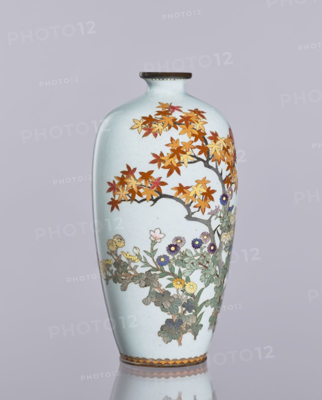 Vase, cloisonné enamel, ovoid elongated vase with a narrow spout; decoration: chrysanthemums, maples, and plant ornaments flowers iconography; Japanese art, Meiji 1868–1912 / Taisho 1912–1926, ca. 1910–1920; maker, Nagoya, Japan; height 12.4 cm, diameter 16.3 cm., A tall, slender vase with a pale bluish-white background featuring painted branches with autumnal leaves in shades of orange, red, and yellow, and a lower cluster of flowers and foliage in greens with blooms in purple, yellow, and pale pink; the vase has a narrow neck with a dark brown rim and a decorative band near the base in brown and golden tones, all shown against a muted light gray-lavender background.
