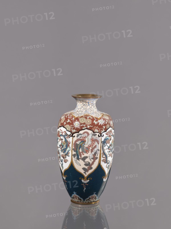 Vase, gift, repossession — phoenixes iconography, plant ornament, dragons iconography; Japanese art, Japanese culture; Japanese dragon motif, hoo phoenix; Meiji 1868–1912; cloisonné enamel. Description: Vase on a small ring-shaped foot with a short neck ending in a spout widening toward the top. Decoration: stylized plant ornament in six reserves alternating dragon and phoenix. Date: ca. 1890–1900 Meiji period. Place of origin: Kyoto, Japan. Medium: cloisonné enamel. Dimensions: height 15.0 cm; diameter 8.0 cm., A decorative vase with a rounded body and narrow neck sits against a plain gray background; its colors include deep blue, rusty red, white, gold, teal, and touches of black and brown, with ornate painted panels showing stylized birds and floral motifs in shades of teal, red, and cream, gold outlining and accents, a band of floral pattern near the shoulder in cream and gold against a red field, and a gold rim at the mouth, with a subtle reflection of the vase visible on the surf...