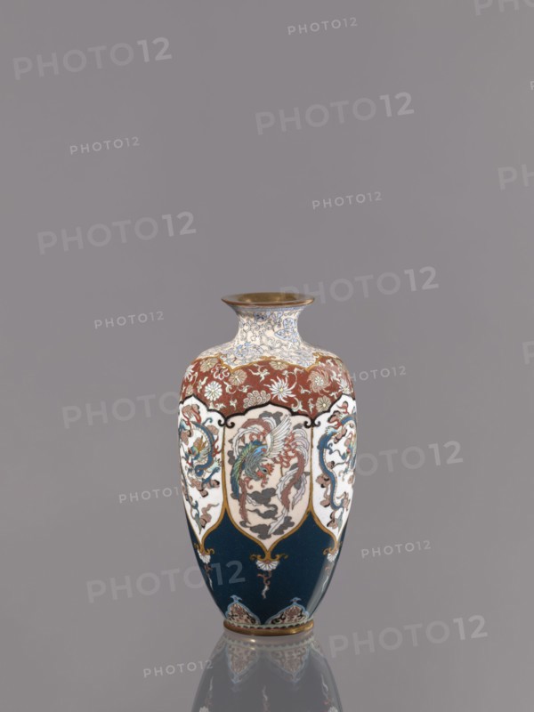 Vase, cloisonné enamel. Vase on a small, ring-shaped foot with a short neck ending in a spout widening toward the top. Decoration: stylized plant ornament in six reserves alternating dragon and phoenix motifs Japanese dragon, hoo/phoenix. Meiji period, ca. 1890–1900. Japan, Kyoto. Vessel; height 15.0 cm, diameter 8.0 cm., A decorative vase with a flared rim and rounded body sits centered against a plain gray background, displaying a color palette of deep blue, white, red, gold, green, and brown. The upper neck area features an intricate white and blue scrolling pattern with gold accents and a metallic gold rim. Below that a band of red with floral motifs in white and gold encircles the shoulder. Panels of white panels framed with gold and black outlines descend along the body, each panel containing ornate multicolored designs including stylized birds and curling foliage in greens, reds, blues, and browns. The lower portion of the vase is a deep blue with small decorative motifs in gold...