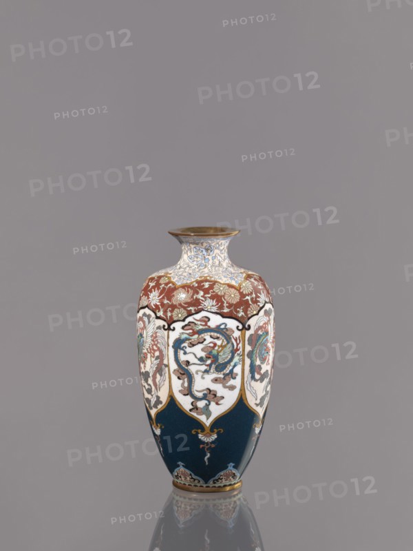 Vase, gift; repossession Kraków. Cloisonné enamel vase on a small ring-shaped foot with a short neck ending in a spout widening toward the top. Decoration: stylized plant ornament in six reserves alternating dragon and phoenix Japanese dragon motif, hoo/phoenix, Meiji period, ca. 1890–1900. Japan, Kyoto. Vessel. Height 15.0 cm, diameter 8.0 cm., A single vase sits centered against a plain gray background, with a rounded body that tapers to a narrow neck and flared rim; the vase displays a palette of deep blue, white, red, gold, and touches of light blue and green, with ornate painted panels featuring flowing, curving motifs and stylized creatures, floral patterns along the shoulder in red and gold, decorative gold outlines separating the panels, and a reflective surface that creates a faint mirrored reflection on the surface below.