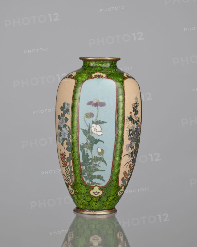 Vase, cloisonné enamel, floral pattern with butterflies and plant ornaments; Meiji period 1868–1912, Japan; background of decoration in reserves: dense green floral rosettes and butterflies; 19th/20th century; vessel; height 15.3 cm, width 8.7 cm, depth 8.2 cm., A tall vase with an elongated ovoid shape standing on a short foot and a narrow neck, decorated with a repeating panel design; the main background color around the panels is a bright mottled green, the panels themselves alternate between a pale beige and a soft blue, and the neck rim and foot edge show a metallic bronze tone. The panels contain floral motifs rendered in greens, whites, purples, blues, reds, and muted pinks, with leafy stems and clusters of blossoms; thin brown outlines separate the panels from the green ground and small decorative cartouche shapes in cream and brown accent the panel borders. The surface has a glossy finish that produces a subtle reflection of the vase on the smooth gray surface beneath it, and ...