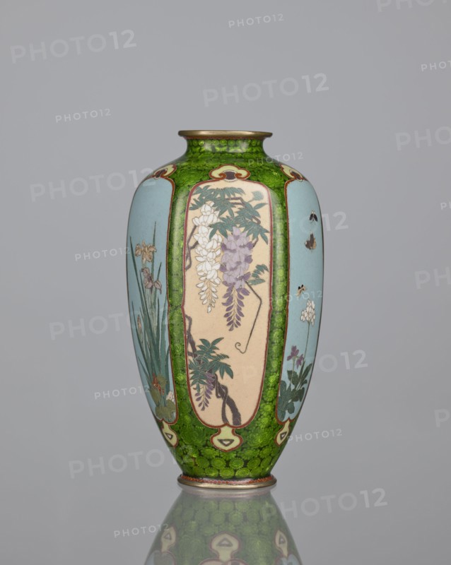 Vase, cloisonné enamel, floral pattern with butterflies and plant ornaments; background of decoration in reserves: dense green floral rosettes and butterflies. Japanese, Meiji period 1868–1912, 19th/20th century. Vessel. Height 15.3 cm, width 8.7 cm, depth 8.2 cm., A tall, narrow vase with a rounded body and a slightly flared neck standing on a reflective surface, decorated with panels framed by a textured green background; the vase shows painted floral scenes in muted tones including light blue, pale beige, purple, white, green, and touches of brown and gold, with leafy branches, hanging blossoms, slender stems, and small insects depicted within the panels, and the green area showing a mottled pattern that continues down to a decorative base ring.