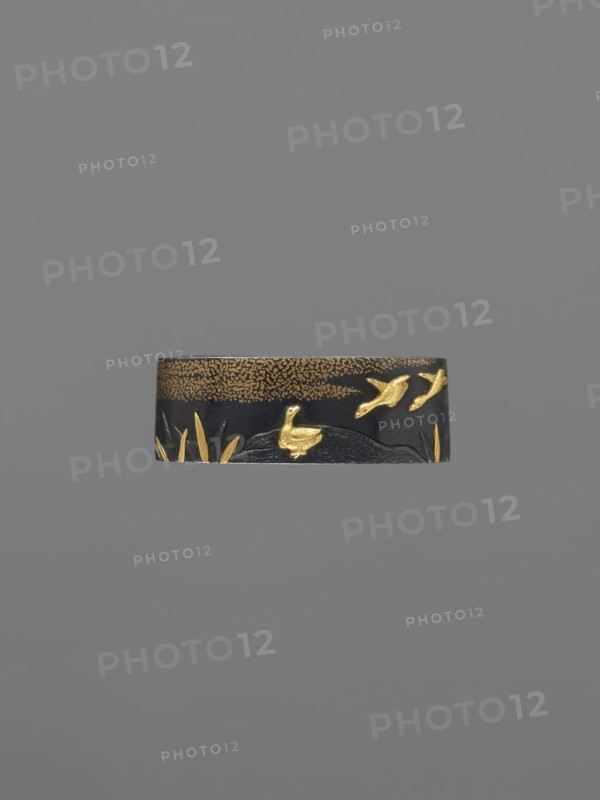 Metal collar ending a sword hilt fuchi, Nara, Toshinaga 1667–1736, Nara school, geese; Plastic decoration: wild geese among the clouds; 18th century Edo period, 1603–1868, Japan; katana hilt component fuchi; height 3.9 cm, width 2.2 cm, depth 1.2 cm., A narrow horizontal band with a dark base color decorated with raised golden elements depicting a group of birds in various poses above a textured area and a few stylized plant shapes, set against a uniform gray background; visible colors include dark blackish tones, gold, and shades of gray.