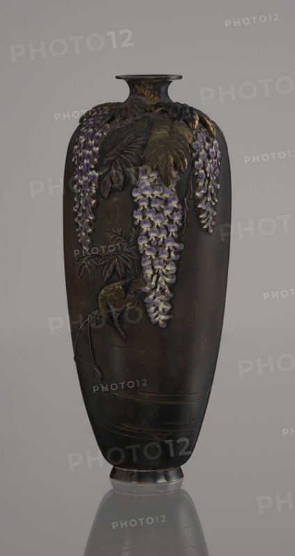 Vase, Toshitsugu maker, Meiji period 19th/20th century, Japan. Ovoid vase on a low ring foot with a very short neck and a strongly flaring, plate-like spout. Decorated in a naturalistic, asymmetrical composition showing a wisteria vine with three clusters of flowers hanging from the top near the neck; a kingfisher perches on a branch near the lowest flower, gazing into engraved water with swimming fish. Decoration executed with ro-iro zogan inlaid color painting combining inlaid metals and alloys with colored enamels white, several shades of lilac, subtle green accents. Leaves and bird figurine are gilded and patinated with shakudo. Lower belly bears engraved inscription: Sumisuiin Toshitsugu sei. Cloisonné enamel vessel, height 16.2 cm, diameter 7.1 cm., A tall dark vase with a narrow neck and flared rim stands against a neutral background, decorated with raised botanical motifs including a few cascading clusters of purple and pale lilac flowers, several elongated leaves rendered in s...