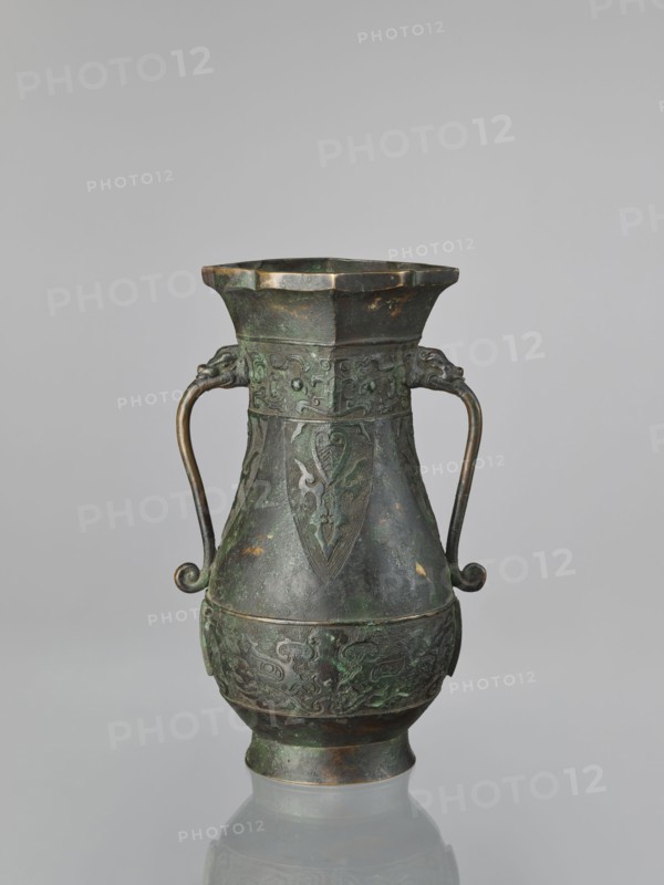 Hu-type vase, hu vessel, Chinese, Ming dynasty 1368–1644, Xuande period 1426–1435, possibly backdated signatures; materials and form: ceramic vessel, ceremonial/ritual hu with taotie ornamentation; dimensions: height 31.5 cm, width 17.0 cm., A single metal vessel with a dark green and brown patina stands against a plain light gray background, featuring a flared octagonal rim, a tapered body with embossed decorative panels, and two curved handles ending in small spirals attached near the upper section; the surface shows areas of darker brown and verdigris green, with intricate raised patterns and a rounded lower body on a slightly flared foot.
