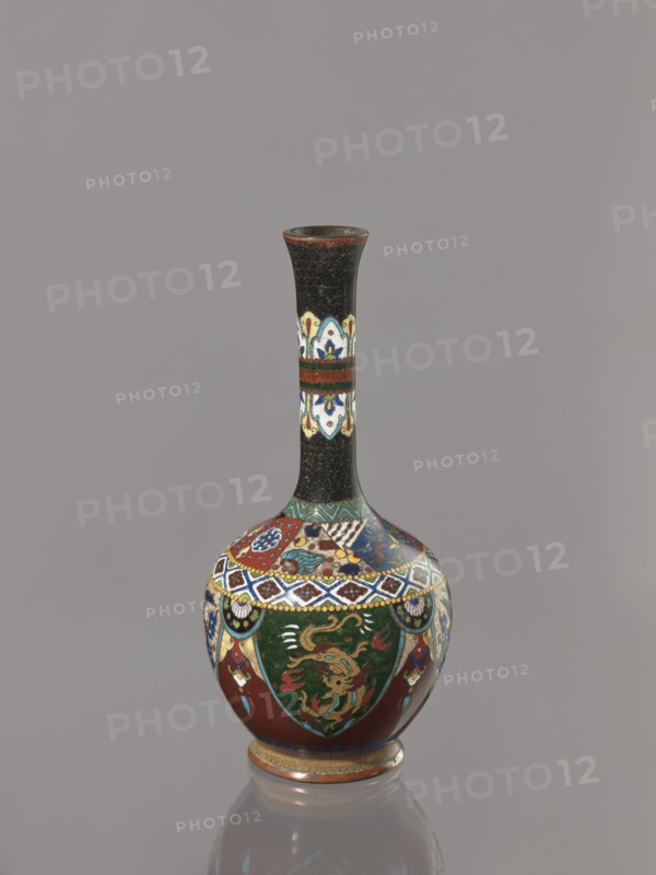 Vase, creator, gift; Japanese art; Meiji period 1868–1912, ca. 1900; Japan. Cloisonné enamel vessel decorated with birds, dragons, and phoenixes. Height 19.3 cm, width 8.9 cm., A tall narrow-necked vase with a bulbous base stands against a plain gray background; the neck is predominantly black with a subtle textured pattern and a horizontal band of decorative panels in white, blue, red, and yellow, while the rounded body features ornate multicolored designs including deep green panels with golden dragon-like figures, red fields, blue and white geometric borders, and accents of yellow and turquoise, all resting on a small decorated foot with reddish-brown and gold tones and showing a reflection on the smooth surface beneath.