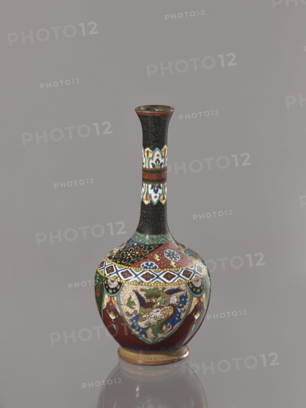Vase, Japanese, Meiji 1868–1912, ca. 1900. Cloisonné enamel vessel decorated with birds, dragons, and phoenixes. Japan. Height 19.3 cm; width 8.9 cm., A tall, narrow-necked vase with a bulbous base sits against a plain gray background; the vase features a dark top section and neck with a textured black pattern, a band of decorative cream, blue, green, and red motifs on the neck, and a wider lower body decorated with intricate patterned panels in red, blue, green, cream, gold, and black, including floral and stylized bird-like designs, geometric borders, and scalloped medallions, with a small stepped foot rim in gold and brown and a faint reflection of the vase visible below.