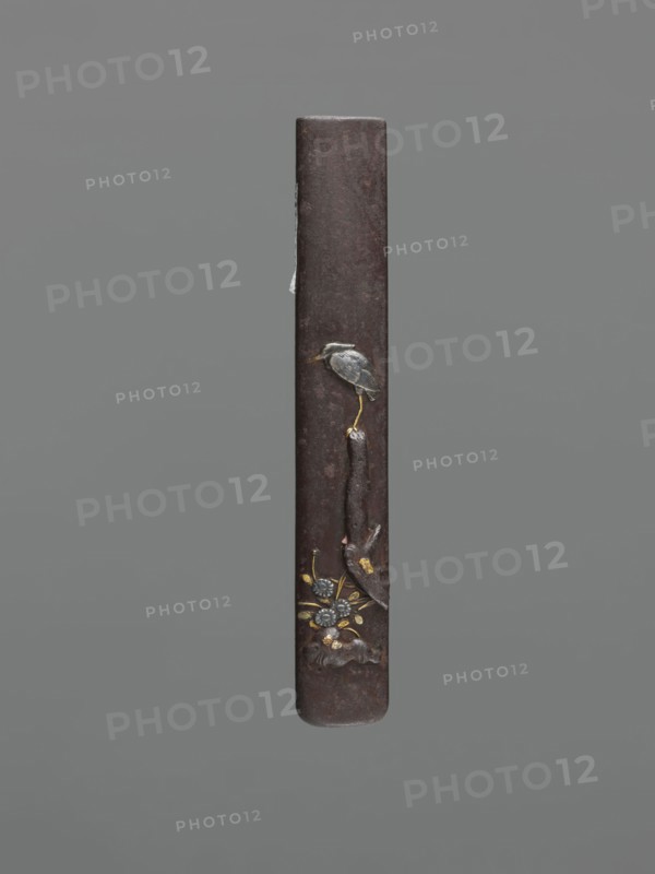 Knife handle kozuka, ; motif: herons; Japanese, Edo period 1603–1868, 18th century; materials/function: kozuka for a katana knife/edged weapon; dimensions: height 9.4 cm, width 1.4 cm, depth 0.6 cm., A slender vertical object with a dark brown, slightly textured surface against a gray background, featuring a small bird figure in gray and muted blue perched on a thin golden leg near the upper middle, a vertical dark brown element extending downward from the bird, and a cluster of decorative floral motifs near the lower section in metallic gold and silvery gray with touches of darker brown.
