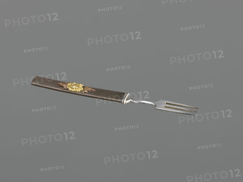 Fork with knife handle kozuka, Krüger, HB dr. Japanese, Edo period 1603–1868, 18th century. Melee weapons / sword accessory kozuka used with katana. Plastic decoration: chrysanthemum flower with two leaves; textured background. Materials: plastic decoration. Function: cutlery/kozuka. Dimensions: height 16.6 cm, width 1.4 cm, depth 0.8 cm., An image of a fork with a long rectangular handle and a three-pronged metal tine section, shown against a solid gray background. The handle appears dark brown with a textured surface and features a decorative floral ornament in gold and bronze tones near one end. The metal tines and connector are silver with some darker discoloration or patina. Overall colors visible include gray, brown, gold, bronze, and silver.