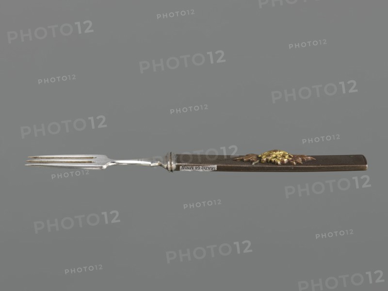 Fork with knife handle kozuka, Japanese, Edo period 1603–1868, 18th century. Melee weapon accessory for swords katana — kozuka. Plastic decoration: chrysanthemum flower with two leaves; textured background. Materials: plastic. Function: cutlery. Dimensions: height 16.6 cm, width 1.4 cm, depth 0.8 cm., A long metal fork with a silver-toned pronged end and a dark brown handle is shown against a solid gray background; the handle features a raised decorative element in gold and copper tones and a silver-toned ferrule where the metal meets the handle.