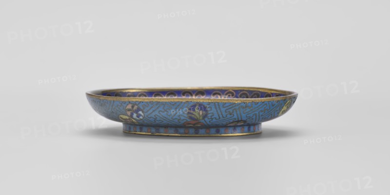 Bowl, gift; Chinese, Qing 1644–1911; cloisonné enamel; 18th century, China; vessel, diameter 9.3 cm. Shallow bowl on a separate foot decorated inside with a colorful composition of three symbolic fruits: a peach, a pomegranate and a finger lemon, accompanied by bats—interpreted as a wish for “many happiness, long life, and many sons.” Inner rim decorated with a circle of ruyi heads black, red, navy blue. Outer surface filled with colorful medallions arranged singly, in pairs, or in threes, featuring stylized plant motifs framed by a Chinese meander pattern made of bent wire in a net-like form on a turquoise background. Foot decorated with a band of alternating red and blue circles on turquoise. Bottom entirely covered with turquoise enamel., A shallow decorative bowl sits on a flat surface, shown in profile with a low rounded rim and a narrow foot. The exterior and interior surfaces display intricate enamel patterns in blue tones, with accents of gold around the rim and foot. There are...