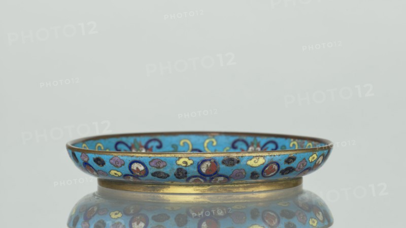 Plate, Chinese, Qing dynasty 1644–1911, 19th century. Cloisonné enamel round plate with a flat bottom, arched rim, on a low foot. Mirror decorated with a tondo surrounded by a multicolored lotus tendril; tondo depicts a cricket in motion perching on flowering plants, positioned at right, with flowering plants dominating the composition. Outer rim filled with medallions showing stylized Chinese symbols lotus, peach surrounded by cloud-like projections. Underside of the bottom covered with single-color turquoise enamel. Vessel; height 2 cm, diameter 14.7 cm., A shallow decorative bowl with a rounded rim sits on a reflective surface, showing a reflection beneath. The bowl has a bright blue background with patterns in colors including gold, yellow, red, purple, black, and dark blue, and a metallic gold band along its base and rim. The design includes small circular motifs and stylized cloud-like shapes in the various colors, and the interior shows similar blue and gold decorative elements....