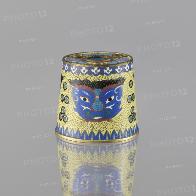 Sawdust container, Cloisonné enamel vessel, Qing dynasty 1644–1911, 19th century, China. Base with four inserts. Decoration on a yellow background: four masks against a grid background; motifs include beasts fantasy, tiger symbol, and meander ornament. Height 8.5 cm; diameter 9.7 cm., A small conical object sits against a plain gray background, its surface decorated with a bright yellow base and intricate patterns in blue, black, red, green, and gold; a stylized blue face with white eyes and red accents is centered on the visible side, flanked by circular green and black medallions, and a band of blue petal-like shapes runs along the lower edge, while a decorative blue and white border encircles the top; the object casts a clear reflection on the glossy surface beneath it.