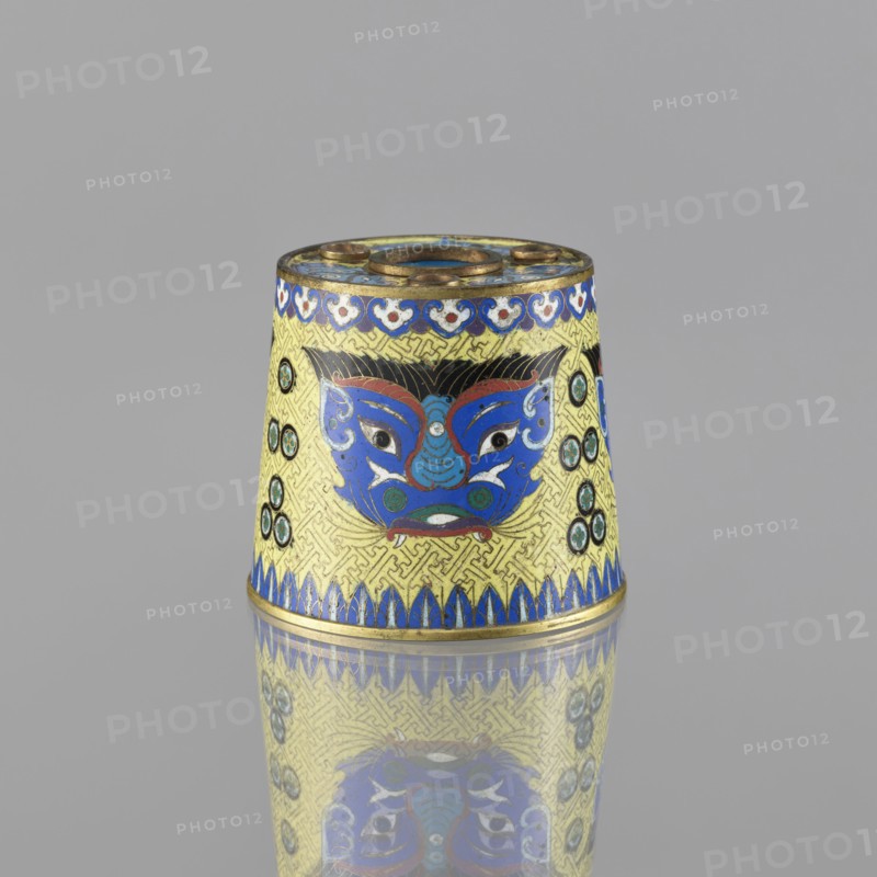 Sawdust container, Paderewski, Ignacy Jan 1860-1941, Chinese art, Chinese culture, Qing 1644–1911, cloisonné enamel, beasts fantasy, tiger symbol, meander ornament. Base with four inserts. Decoration: on a yellow background, four masks against a grid background. 19th century, China. Vessel; height 8.5 cm, diameter 9.7 cm., A conical decorative object sits on a reflective surface, showing a reflection below. The object features a dominant yellow background with an intricate repeating pattern and prominent blue, red, white, green, black, and gold decorative elements. A stylized face motif in blue with red and white accents appears on the visible side, flanked by circular green and black roundels on both sides. The top rim and base are accented in gold with blue decorative borders, and the top surface has circular openings and gold detailing. The overall scene includes the gray backdrop behind the object.